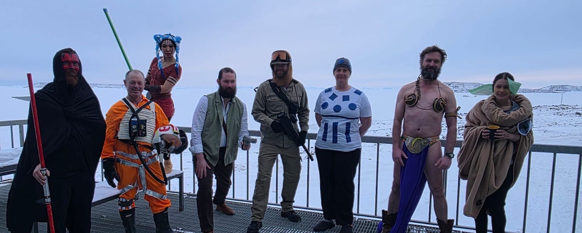 8 expeditioners stand on a metal deck in front of an expanse of sea ice. From left to right they are dressed as the following characters from Star Wars: Darth Maul, X-Wing Fighter Pilot, Padme Amidala, A Jedi, a fighter from Hoth, R2D2, Princess Leia looking surprisingly handsome and Grogu.