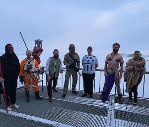 8 expeditioners stand on a metal deck in front of an expanse of sea ice. From left to right they are dressed as the following characters from Star Wars: Darth Maul, X-Wing Fighter Pilot, Padme Amidala, A Jedi, a fighter from Hoth, R2D2, Princess Leia looking surprisingly handsome and Grogu.