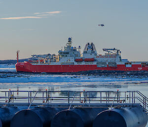 Ship RSV Nuyina in the water in front of Mawson Station, with helicopter overhead and Mawson's bulk fuel farm in immediate foreground