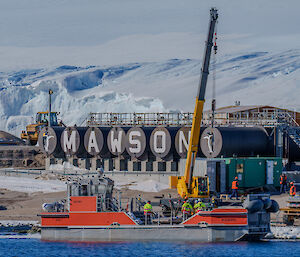 A barge in the water alongside Mawson's wharf, with a crane doing a lift and the Mawson fuel tanks inscribed with teh station's name in the background.