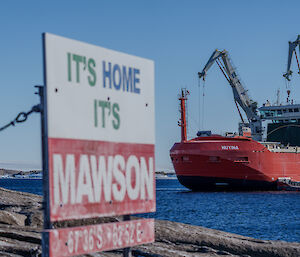 Nuyina in the water, behind the station's "It's Home It's Mawson" sign, doing cargo lifting with gantry cranes and a barge alongside.