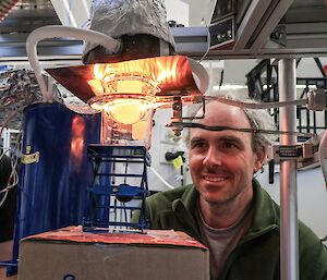 A man looking at a flask containing a small piece of ice. A heat lamp is casting an orange glow across the ice and his face.