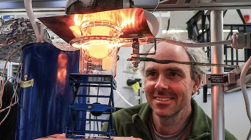 A man looking at a flask containing a small piece of ice. A heat lamp is casting an orange glow across the ice and his face.