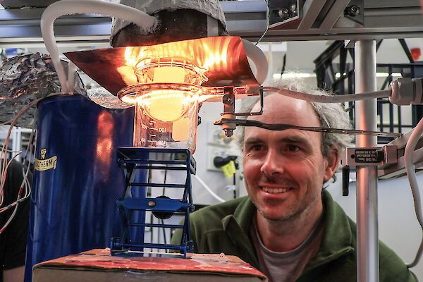 A man looking at a flask containing a small piece of ice. A heat lamp is casting an orange glow across the ice and his face.