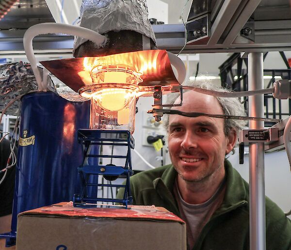 A man looking at a flask containing a small piece of ice. A heat lamp is casting an orange glow across the ice and his face.