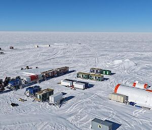 Aerial view of a ice core drilling camp on the Antarctic ice sheet. The camp includes a cylindrical drill tent and about 12 shipping containers housing camp accommodation, living areas and services.