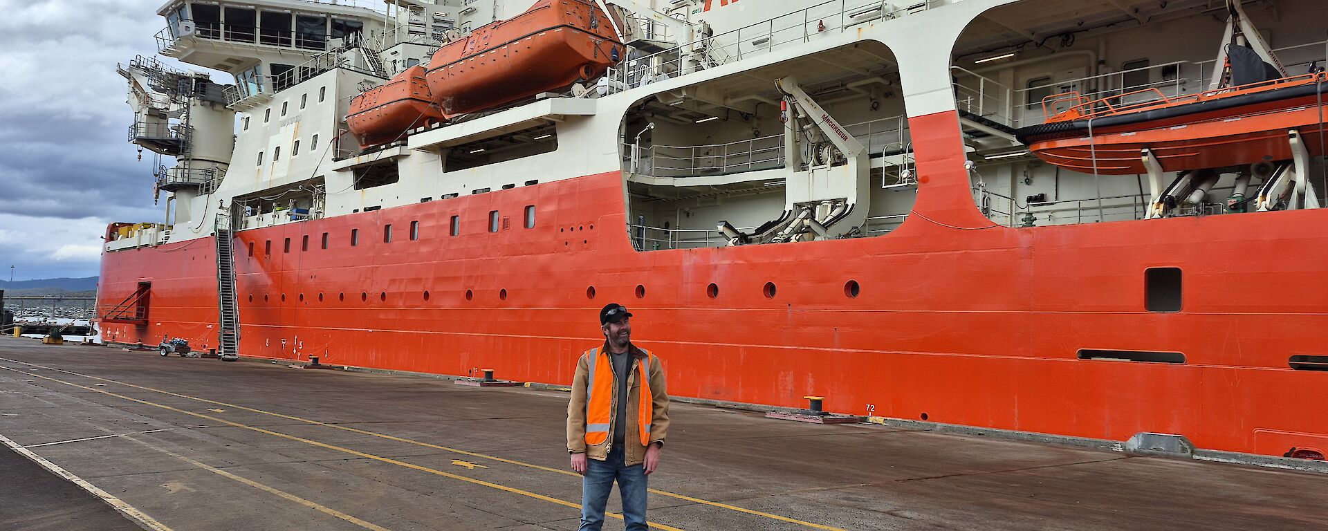 A man wearing jeans and a high visibility vest stands on tarmac in front of a large red and white ship labelled 'Antarctica', with grey cloudy skies overhead.