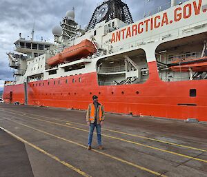 A man wearing jeans and a high visibility vest stands on tarmac in front of a large red and white ship labelled 'Antarctica', with grey cloudy skies overhead.