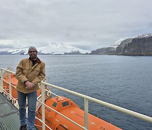 A man wearing blue jeans, a brown jacket and  beanie leans against the side rail of a ship, with orange life rafts behind him. In the background you can see an island with large rocky cliffs and a white mountain.