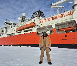 A man wearing brown pants and jacket and a high visibility vest stands on ice in front of a large red and white ship labelled 'Antarctica', with a blue sky dotted with clouds overhead.