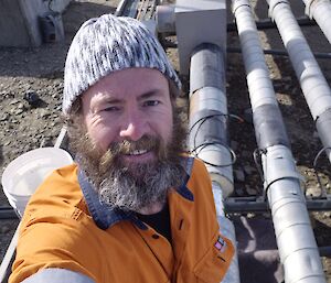 A man wearing a high visibility short and a greay and white beanie poses for a selfie in front of a series of grey pipes running just above a brown rocky ground