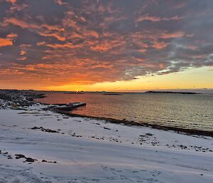 An orange sunset spreads across a sky that is partially covered with fluffy clouds. In the foreground is a snowy beach and calm water