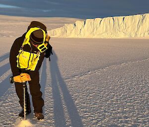 An expeditioner standing on the sea ice, drilling the ice with ice cliffs in the background