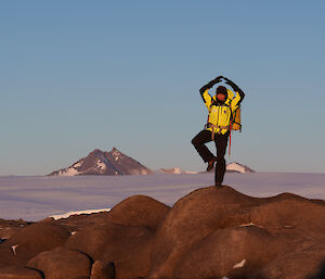 An expeditioner posing on a rocky outcrop, standing on one leg - with Jagged mountain range in far background