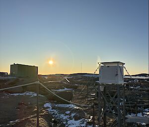 Picture of the Stevenson Screen in foreground and green building in background - with a low horizon sun in the middle