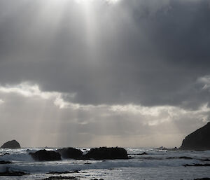 Rays of sunlight filtering through grey clouds on to a grey ocean, that is churning with white surf and bull kelp