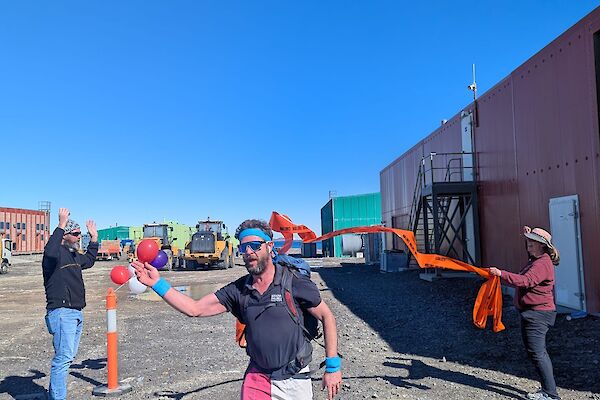 A man wearing pink and white shorts and an electric blue headband to look like a Ken Doll crosses a finish line while others cheer in th ebackground