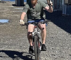 A man with a red beard is riding a bicycle, signalling success to the camera as he rides by