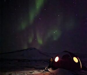 A small red dome, with two windows glowing with light from inside sits in a snowy landscape with a green and purple aurora stretching overhead