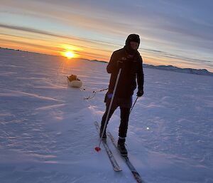 A man wearing black clothes and a balaclava skies towards the camera, towing a sled, with the sun just dipping below the horizon in the background