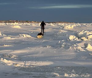 A man skies away from the camera, through a snowy landscape filled with small snow drifts, under a grey, cloudy sky