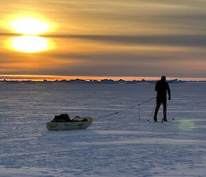 A man skies away from the camera, towing a sled while the sun sets very orange in the background