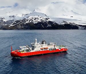 A big red and white ship in a bay with a snow covered volcano behind