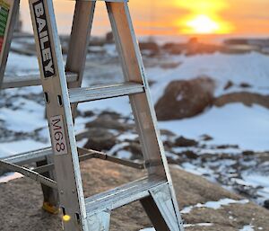 A ladder perched on a rock, with snow covered ground and a sunset in the background