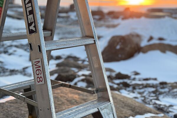 A ladder perched on a rock, with snow covered ground and a sunset in the background