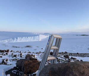A ladder propped up against a large rock, overlooking the sea ice and ice cliffs in the background