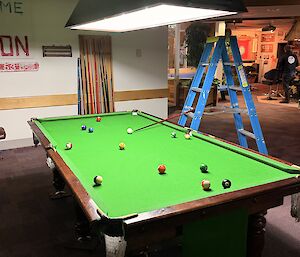 A ladder with a pool cue propped up near a pool table, 'playing' a game of nine ball.