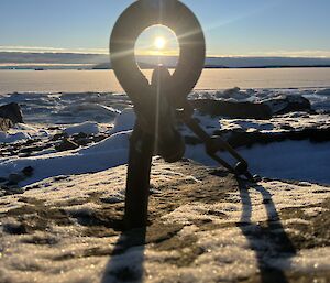 A large metallic eyebolt and chain sticks out of anow. Centred in the circle of the eye is the setting sun un a clear pale blue sky
