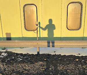 The silhouette of a man against a pale green building, his shadow appears to be holding the shadow of a sensor that is visible in the picture