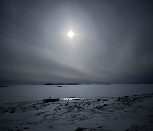A very bright moon glows in a dark sky, lighting up the white sea ice underneath and showing the contrast of rocks in a snowy landscape