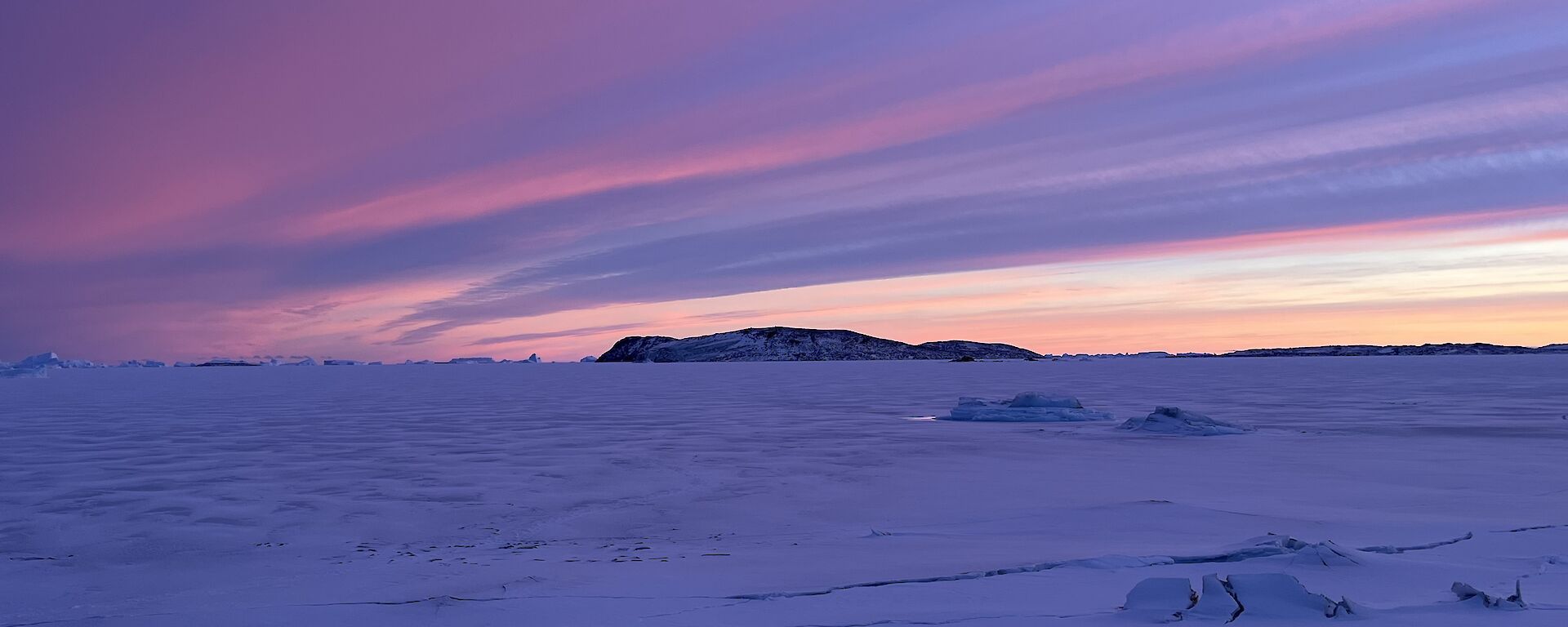 A pink and purple sunset streaks the sky above a snowy landscape, with a rocky outcrop visible in the middle distance