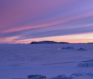 A pink and purple sunset streaks the sky above a snowy landscape, with a rocky outcrop visible in the middle distance