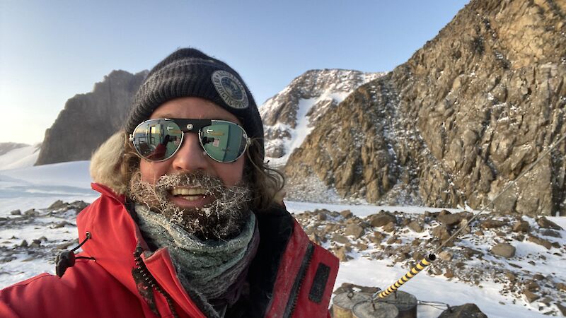 A selfie of a man with ice in his beard and a scenic mountainous backdrop behind.