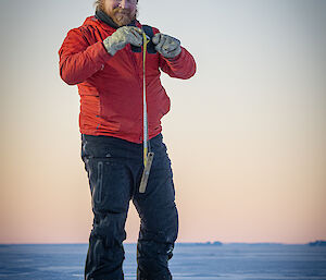 An expeditioner standing on the sea ice, holding a sea ice measuring tape, after having drilled a hole into the ice to measure