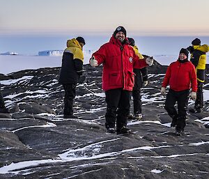 6 expeditioners on an island's rocky outcrop with the sea ice and a tabular ice berg in the distance.