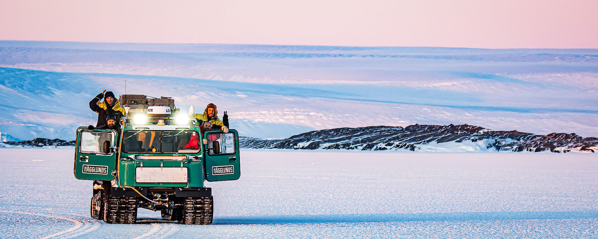 Four expeditioners in an over-snow tracked Hagglands vehicle, waving to the camera while travelling on the sea ice.