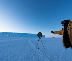 An expeditioner on the sea ice throwing a dart at a dart board mounted on a camera tripod, with tabular iceberg in background
