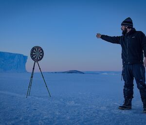 An expeditioner on the sea ice throwing a dart at a dart board mounted on a camera tripod, with tabular iceberg in background