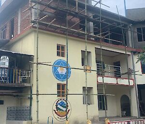 An existing white building in Nepal of two stories has scaffolding to support the construction of two newfloors above