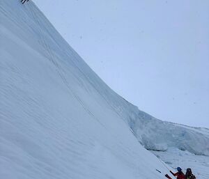 Two people at the base of an ice cliff. A third person on a rope is near the top of the ice cliff.