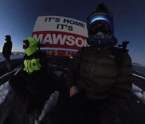 Three expeditioners looking outwards in the early morning light, next to the Mawson sign on West Arm, with the station in the background