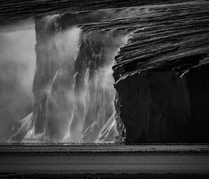 A black and white photo of rugged ice cliffs with snow-blown wind rolling off the cliff-face
