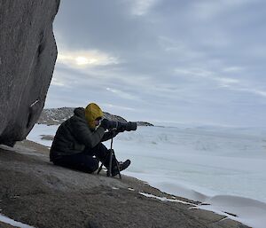 A seated expeditioner looks through a camera mounted on a tripod, waiting to take a photo with ice and snow in background