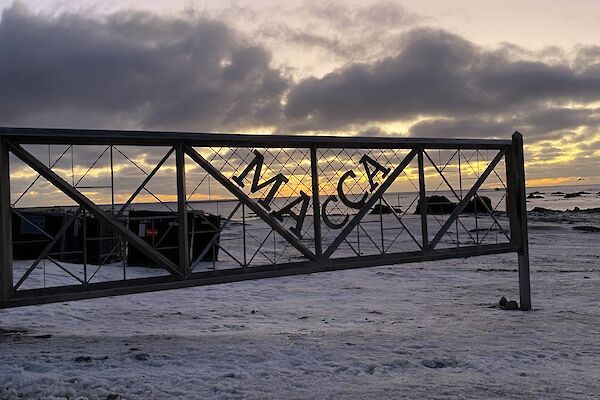 The sunset on a snow-covered beach viewed through a metal gate with the words 'Macca" emblazoned on it.