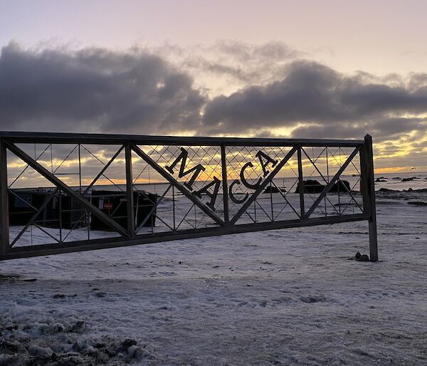 The sunset on a snow-covered beach viewed through a metal gate with the words 'Macca" emblazoned on it.