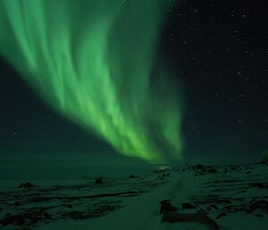 A large and very bright green aurora sweeps down through a dark and starry night, ending at a point above a snowy landscape with a few unusually shaped buildings barely visible in the low light
